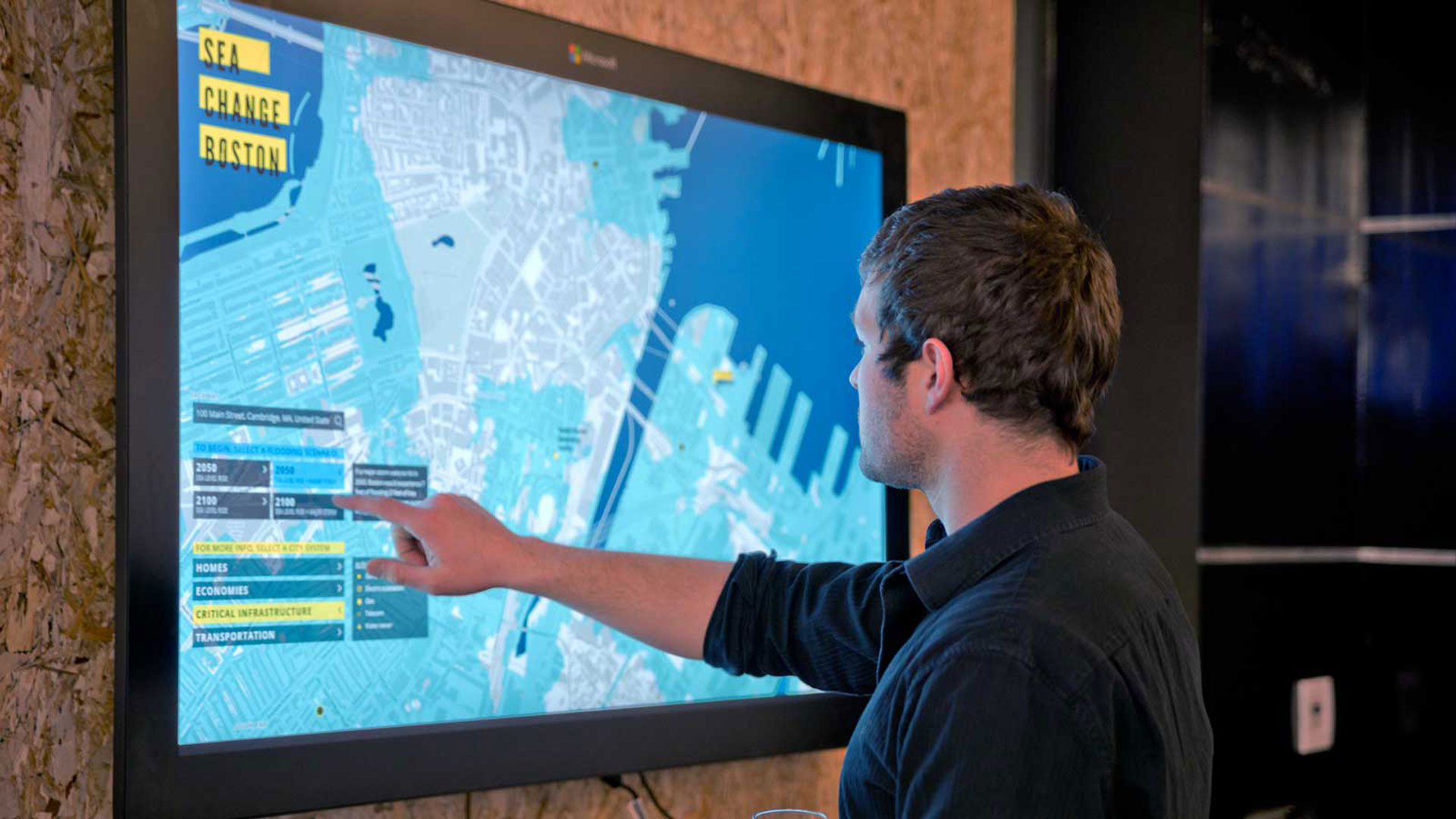 A photograph of a man touching a screen with a map of Boston MA on it. The map is showing flood zones around the city. There is Interactive UI on the screen in the bottom left. Deisngs made with HTML and CSS in collaboration with a developer.